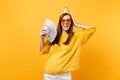 Laughing young woman in orange heart glasses birthday hat putting hand on head holding bundle lots of dollars cash money