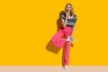 Laughing young woman in pink high waisted baggy pants and sneakers is posing on one leg in front of yellow wall