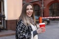 Laughing young woman on a walk in the city drinks coffee from a craft red cup. Beautiful blonde woman in a stylish coat on a sunny Royalty Free Stock Photo