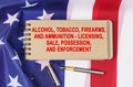 Against the background of the flag of the USA lies a notebook with the inscription - ALCOHOL, TOBACCO, FIREARMS