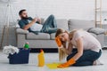 Woman suffers from headache and does cleaning at home, man with beard lies on couch and plays on phone Royalty Free Stock Photo