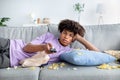 Lazy black teen guy lying on couch with scattered snacks, holding remote control, watching TV at home Royalty Free Stock Photo