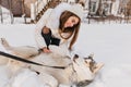 Lazy dog lying on the snow while beautiful girl in woolen hat sitting beside. Outdoor portrait of excited woman in white Royalty Free Stock Photo