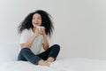 Lazy morning and bedtime concept. Positive Afro woman holds mug of hot tea or coffee, sits in bed, looks gladfully aside, enjoys