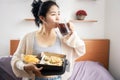 Lazy Asian woman eating junk food hand holding box of pizza and bowl of potato chips drinking sweet soda in bed