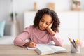 Learning concept. Tired and bored african american girl sitting at desk at home, writing in notebook, doing homework Royalty Free Stock Photo