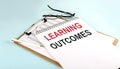 LEARNING OUTCOMES text on notepad on clipboard with chart on blue background, concept closeup. Business and finance concept Royalty Free Stock Photo