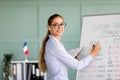 Lecture concept. Millennial female French teacher standing near whiteboard, writing grammar rules and smiling at camera