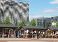 Leeds outdoor market with people shopping at traditional stalls selling clothing and food in front of the modern victoria quarter