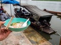 Left hand holding a bowl of noodles. The river is behind and there are noodle boats. thailand lifestyle
