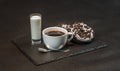 left view on an aromatic black coffee in a white cup with coconut liqueur in a glass, with two oreo doughnut on a black stone