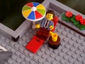 A Lego businessman minifigure relaxing on a sun lounger in the shade of a colorful parasol that is on a rooftop