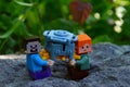 LEGO Minecraft figures of Steve and Alex sitting on garden rock in shadow of garden plants, holding large toy money safe.