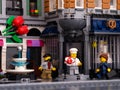 A Lego pastry chef with a cake standing on the doorstep of a his bakery, painter and businessman minifigutes walking near