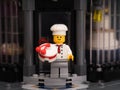 A Lego pastry chef minifigure with a cake standing on the doorstep of a bakery