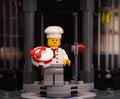 A Lego pastry chef minifigure with a cake standing on the doorstep of a his bakery