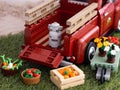 A Lego Pickup Truck with milk bucket in its bed, some crates of vegetables and a wheelbarrow with flowers near it on grass