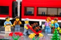 Lego porter with stack of suitcases and some passengers on the platform in front of a red train