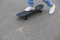 The legs of a child in jeans and white sneakers stand on a skateboard on an asphalt road. A child rides a skateboard on