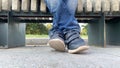 Legs feet of a bored teen boy sitting on a bench in urban city park and wearing jeans and shoes, he is lazy and crossing his legs