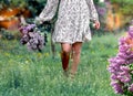 Legs of a girl in a light dress walking on green grass with a bouquet of lilacs. Background - nature, sun, rays, bokeh effect