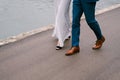 Legs of the groom in blue trousers and the bride in a white dress are walking along the asphalt road over the sea. Close
