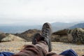 Legs of a man resting in boots for mountain tracking against the backdrop of mountains and valleys with noisy clouds