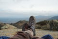 Legs of a man resting in boots for mountain tracking against the backdrop of mountains and valleys with noisy clouds