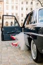 Legs of pretty bride woman in red high-heals sitting in black retro car, old buildings of the city on the background.