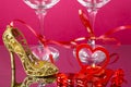 Legs of two champagne glasses with red ribbons on a pink background next to a heart candlestick with a candle and a