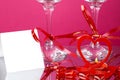 Legs of two champagne glasses with ribbons on a pink background and and a card and a heart candlestick with a burning candle