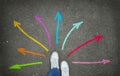 Legs in white sneakers in front of colorful arrows pointing in different directions on the gray asphalt