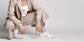 Legs of woman in beige business smart casual suit and sneakers sitting over light background. Stylish business female