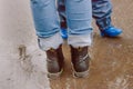 The legs of a young woman in blue jeans, and the legs of a child in rubber boots stand in a puddle on the asphalt