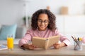 Leisure concept. Cute black schoolgirl reading paper book and drinking orange juice, sitting at desk Royalty Free Stock Photo