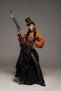 Length view of attractive steampunk woman in top hat holding gun and standing with hand on hip on grey Royalty Free Stock Photo