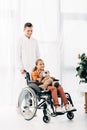 Length view of pediatrist in white coat and kid with teddy bear on wheelchair