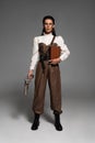 Length view of steampunk woman holding book and vintage pistol on grey