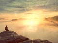 Tourist sit on peak of sandstone rock and watching into colorful mist and fog in morning valley. Sad man. Man sit. Man in jeans. Royalty Free Stock Photo