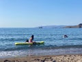 Lesvos, Greece - 29 August 2020: Attractive young woman wearing a pink bikini sitting relaxing with friend on sup board Royalty Free Stock Photo