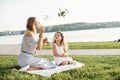 Let the launch of bubbles has come. Photo of young mother and her daughter having good time on the green grass with lake