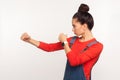 Let`s fight! Side view of determined confident girl with hair bun in denim overalls punching, boxing with clenched fists, struggl