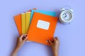 Let`s go back to school: the child`s hands hold an orange notepad, next to a white alarm clock, purple background, top view, Royalty Free Stock Photo