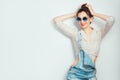 Lets dance! Young beautiful cheerful woman with hair bun in sunglasses wearing in jeans shorts dancing and posing over white wall.