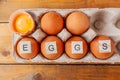 Lettering in wooden square letters eggs on brown eggs in cardboard box with empty cells and one broken on brown wooden table, top Royalty Free Stock Photo