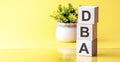 letters of the alphabet of dba on wooden cubes, green plant on a yellow background. dba - short for Data Base