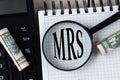 Letters MRS under a magnifying glass on a white notebook against the background of a fragment of a calculator and banknotes