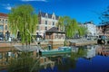 View over town moat on old medieval buildings, ancient eel fishing boat, quay with waterfront restaurant, green trees, clear blue