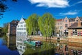 View over town moat on old medieval buildings, ancient eel fishing boat, quay with waterfront restaurant, green trees, clear blue