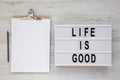 `Life is good` words on a lightbox, clipboard with blank sheet of paper on a white wooden surface, top view. Overhead, from abov Royalty Free Stock Photo